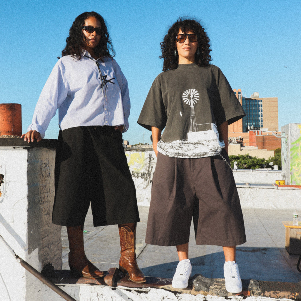 Two models standing on a rooftop ledge under a clear blue sky, showcasing streetwear outfits. The model on the left wears a light blue button-down shirt, black wide-leg shorts, and brown boots.