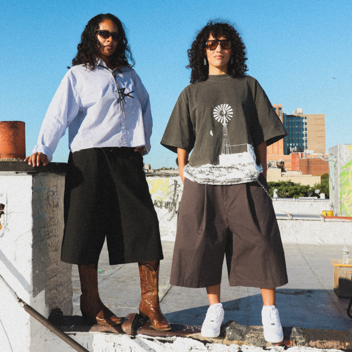 Two models standing on a rooftop ledge under a clear blue sky, showcasing streetwear outfits. The model on the left wears a light blue button-down shirt, black wide-leg shorts, and brown boots.