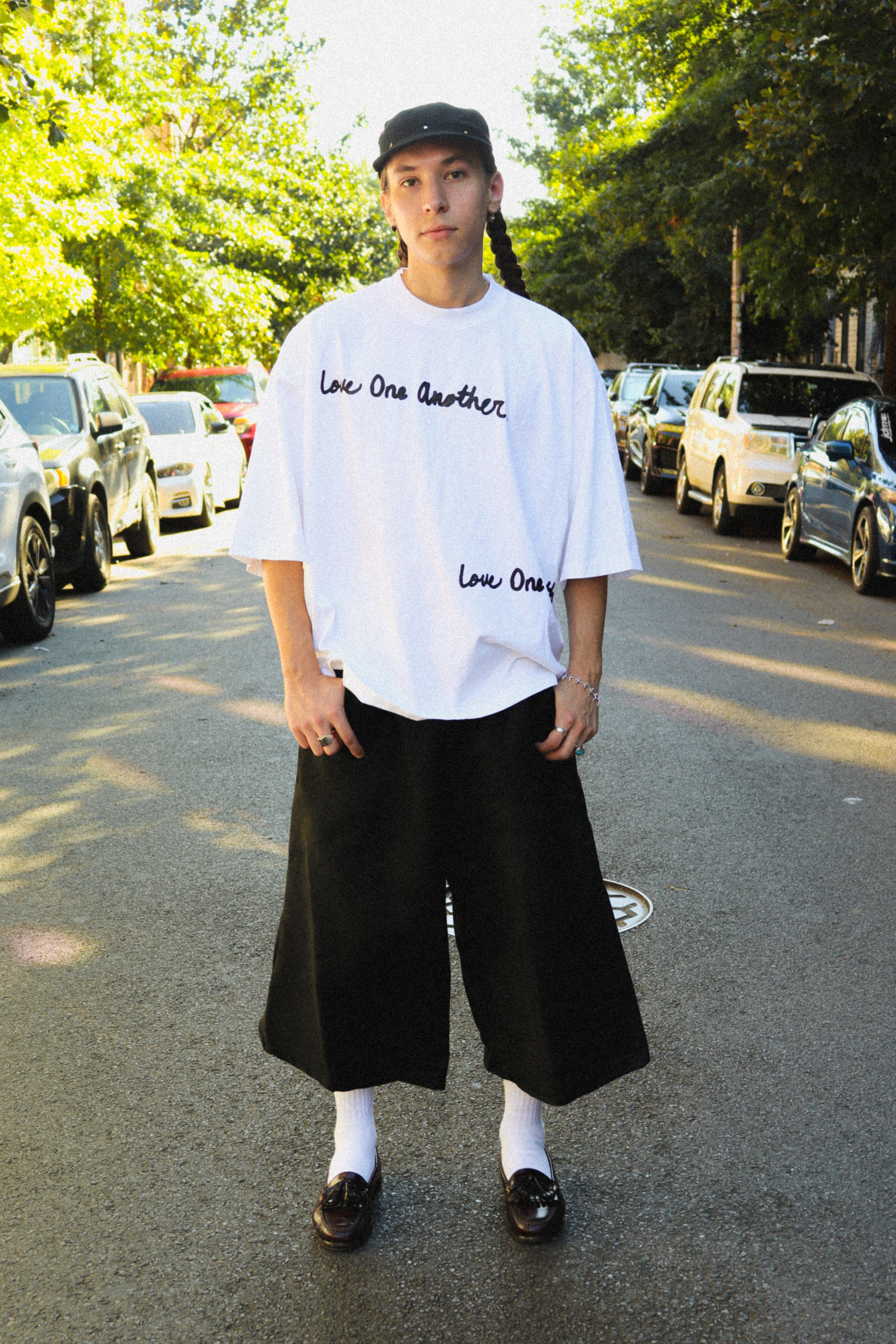 A person standing on a sunlit street wearing an oversized white T-shirt with black script text, loose black wide-leg pants, white socks, and dark loafers.