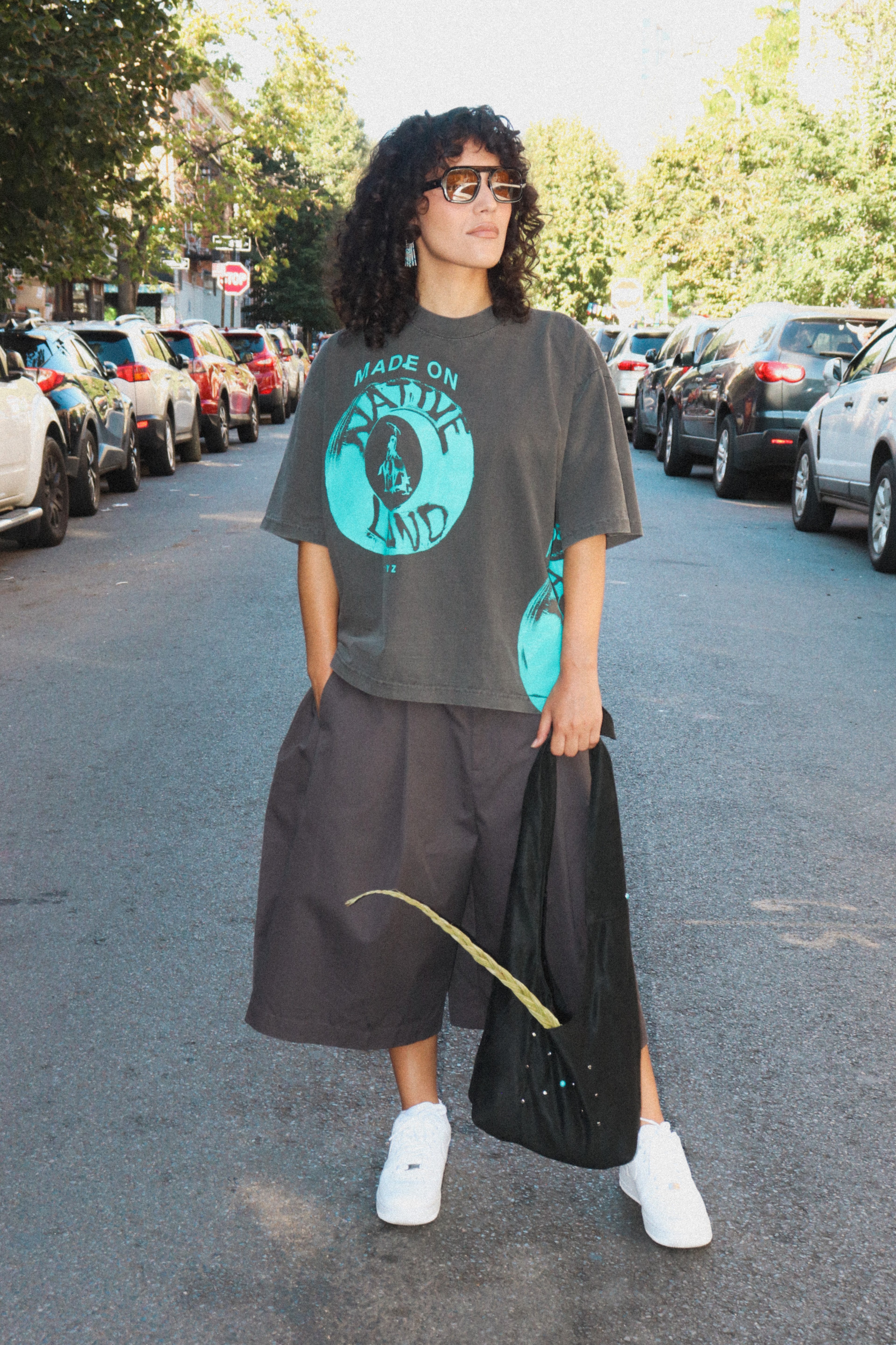A person standing on a street wearing an oversized dark gray graphic T-shirt with a bright teal circular print, dark wide-leg shorts, white sneakers, and holding a black tote bag.