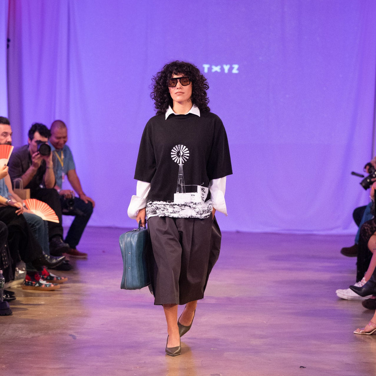 A model walks down a runway wearing a black graphic t-shirt with a white collar, dark wide-leg cropped pants, and carrying a teal bag. The background is a purple-lit runway show with audience members seated on both sides.