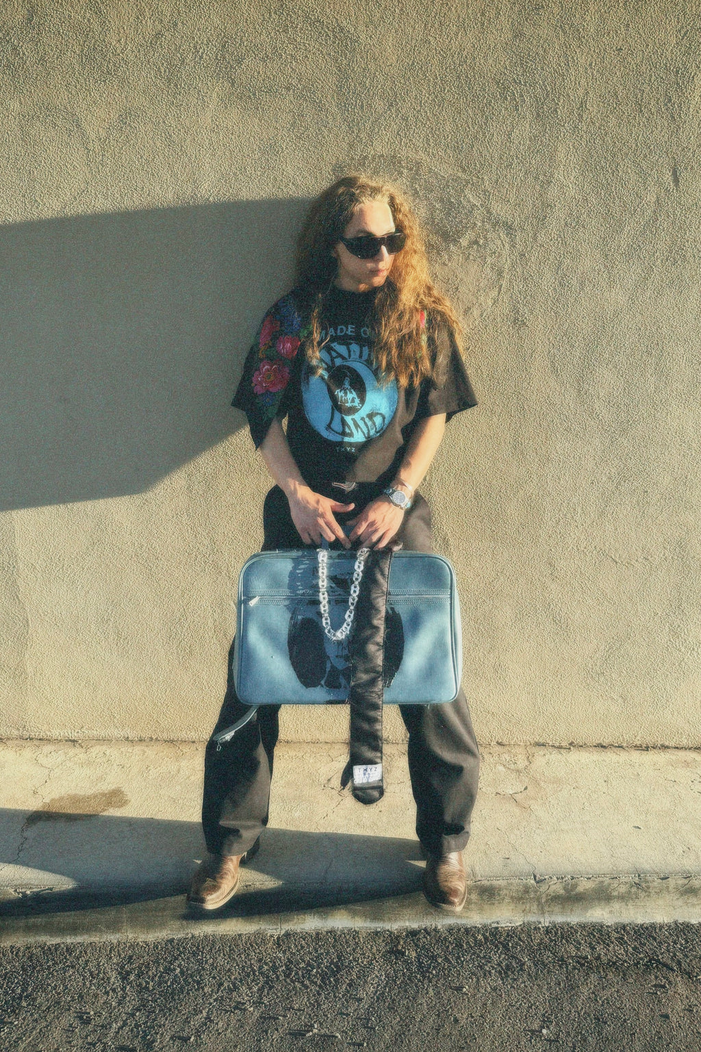 Person with long hair and sunglasses holding a suitcase against a concrete wall.