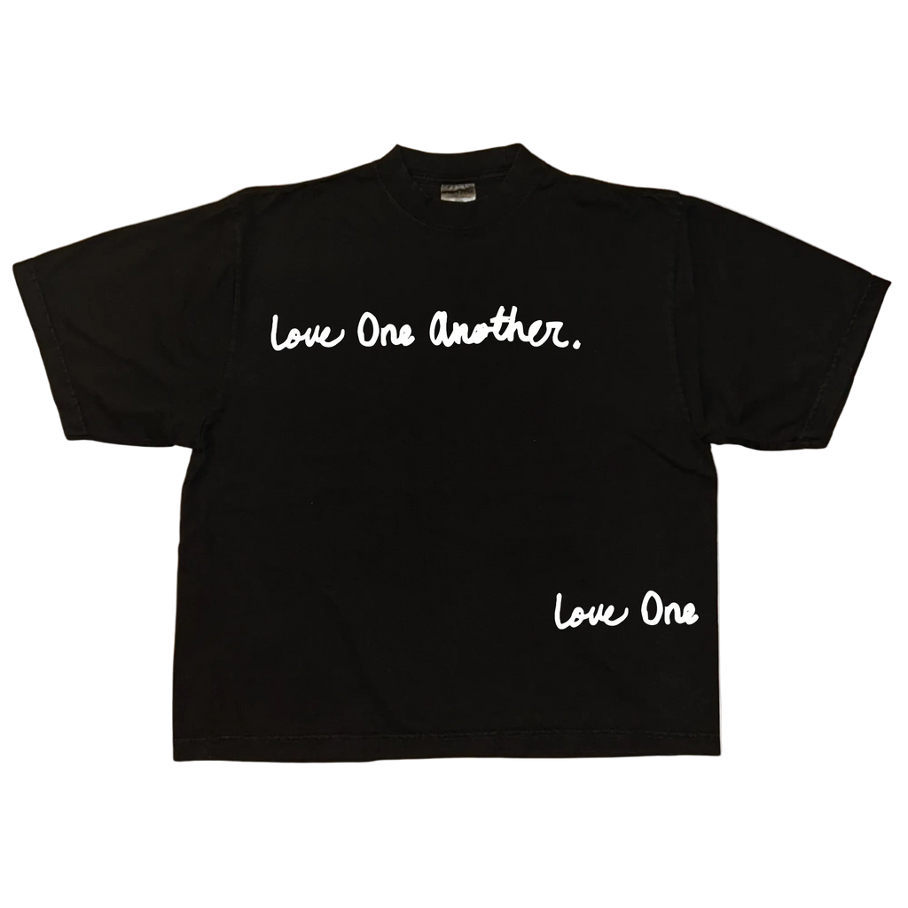 A black short-sleeve t-shirt with white script text reading 'Love One Another.' across the chest and 'Love One' near the lower right hem, displayed flat against a white background.